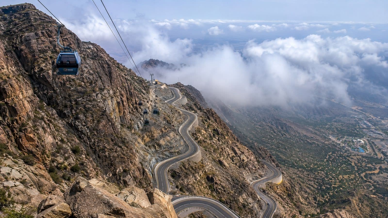 Panoramic view of Al Taif’s mountain road, cable cars, and cloud-covered highlands.