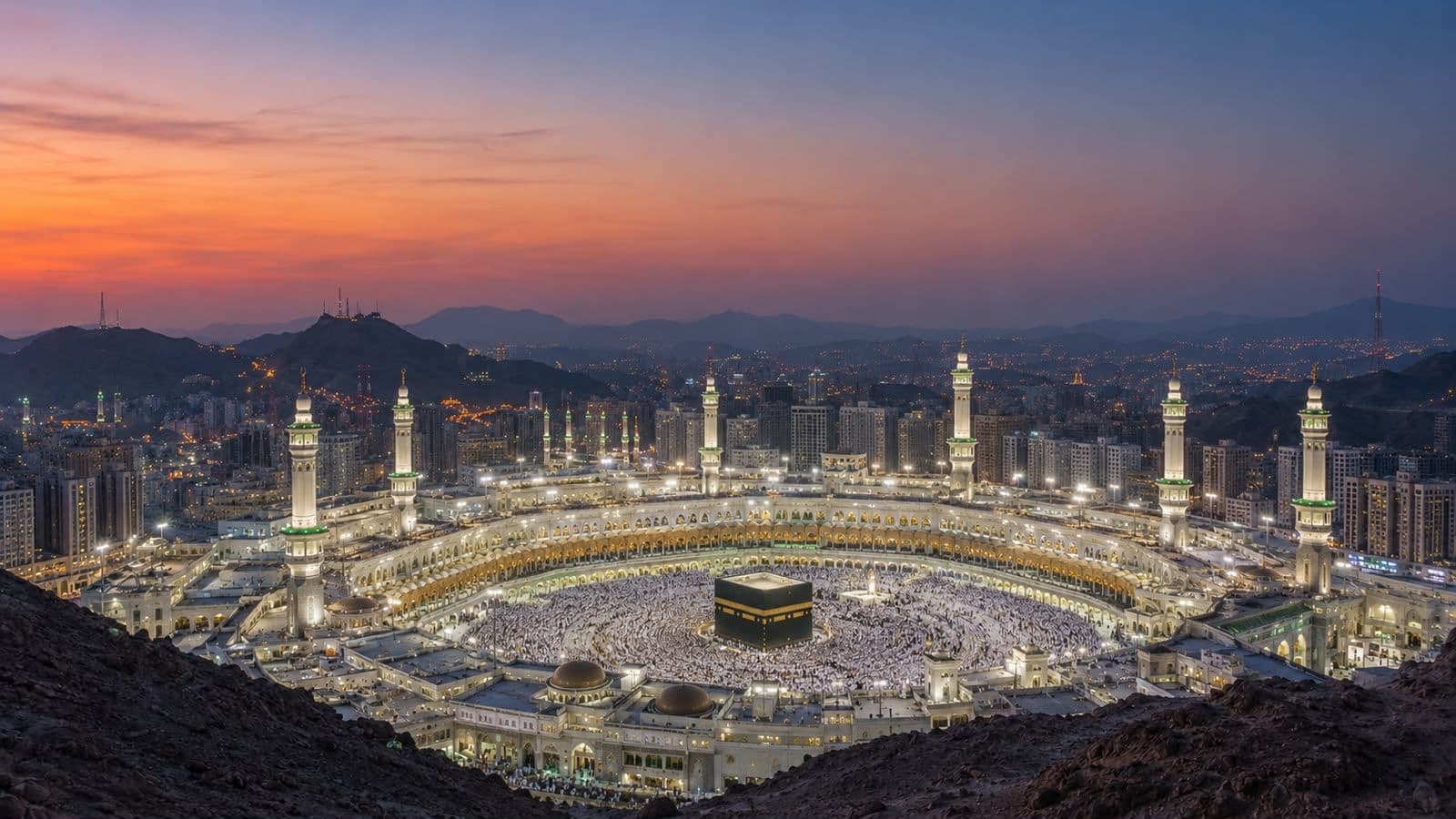 Aerial view of Masjid al-Haram in Makkah at sunset, showcasing the Kaaba surrounded by worshippers and illuminated minarets against a mountainous skyline.