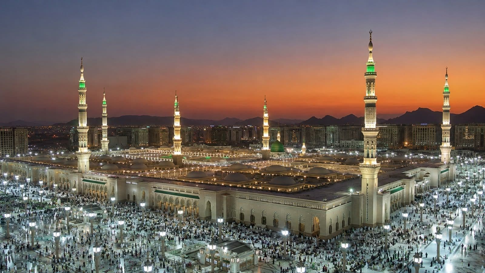 Al-Masjid an-Nabawi in Madinah at sunset, featuring its iconic green dome, illuminated minarets, and expansive courtyards.